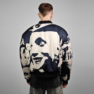 May include: A black and cream-colored bomber jacket featuring a large, stylized face print. The jacket has a quilted design and a black collar and cuffs. The back view shows the full graphic, creating a bold statement piece.