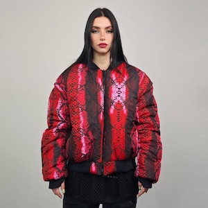 Op de afbeelding: Een rode en roze bomberjack met een slangenprint en een ritssluiting. De jas heeft een zwarte geribbelde kraag en manchetten.