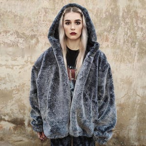 Pode incluir: Casaco com capuz de pele sintética cinzenta com padrão de pele de cobra. O casaco tem um corte folgado e um capuz grande. A modelo está a usar uma t-shirt preta. É uma peça de destaque.