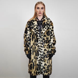 May include: A long leopard print coat with black and tan spots. The faux fur coat has a wide collar and long sleeves. The coat is worn over a black top and black gloves. The model is standing in front of a white background.