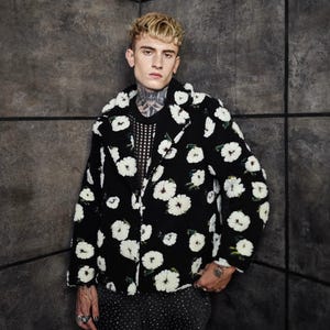May include: A black jacket with a white floral pattern. The jacket has a classic lapel and is worn over a black, textured top. The person is wearing black and white patterned pants. The background is a dark gray wall.