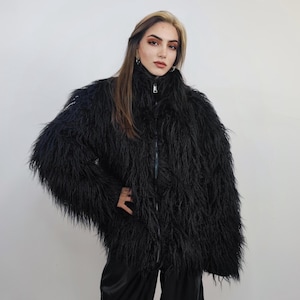 May include: A black faux fur jacket with a high collar and a full-length zipper. The jacket has long, shaggy fur and a relaxed fit. The model is wearing black pants. The background is a plain white wall.