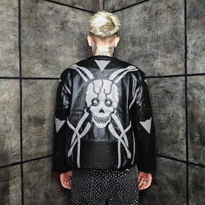 Peut inclure: Veste en cuir noire avec un grand motif de crâne et d'os blancs dans le dos. La veste a une taille côtelée et des accents blancs sur les épaules. La veste est portée par une personne avec une coiffure blonde.