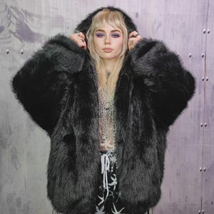 Könnte beinhalten: Schwarze Kapuzenjacke aus Kunstfell mit Reißverschluss. Die Jacke hat ein weiches, strukturiertes Aussehen. Das Model trägt die Jacke mit einem durchsichtigen Oberteil und einer gemusterten Hose. Der Hintergrund ist eine strukturierte graue Wand.