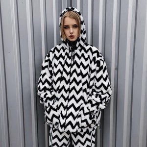 Op de afbeelding: Een zwart-witte chevron-gepatroneerde capuchontrui en bijpassende broek. De trui heeft een ritssluiting en de capuchon is gevoerd met wit bont.