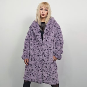 May include: A long, lavender faux fur coat with a large collar and a textured pattern. The coat is knee-length and has a relaxed fit. The coat is worn over a black top and black leggings.