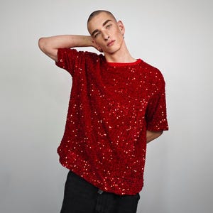 Può includere: Una maglietta rossa con paillettes e girocollo rosso. La maglietta a maniche corte è ricoperta di piccole paillettes lucenti. La maglietta è abbinata a pantaloni neri. L'insieme è audace e accattivante.