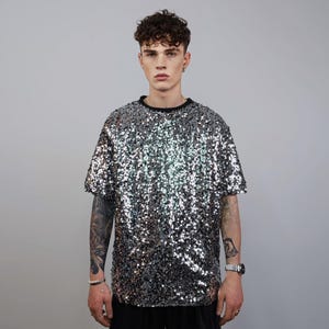 May include: A silver sequin t-shirt with a black crew neck. The short-sleeved top is covered in shiny, reflective sequins. The shirt is a statement piece, perfect for parties or special events.