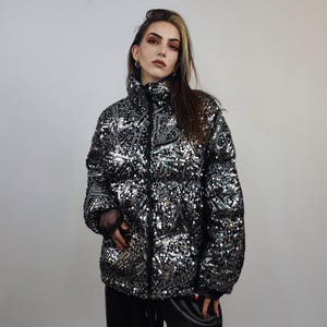May include: A black puffer jacket covered in silver sequins. The jacket has a high collar and a full-length zipper. The sleeves are long, and the jacket appears to be warm and stylish. The jacket is a statement piece.