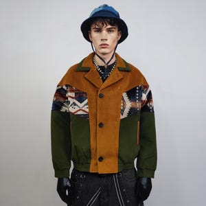 Puede incluir: Chaqueta marrón, verde y estampada con cierre de botones. La chaqueta tiene una textura de pana y presenta un diseño contrastante en los hombros. Un sombrero de pescador azul completa el look.