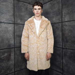 May include: A long, beige faux fur coat with a spotted pattern. The coat has a notched collar and a button closure. The model is wearing a white shirt underneath. The background is a dark gray wall with a grid pattern.