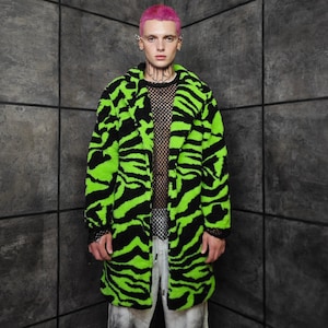 May include: A long, vibrant coat with a bold zebra stripe pattern in neon green and black. The coat has a plush, textured appearance and a classic lapel design. The model is wearing a black mesh top and light-colored pants.