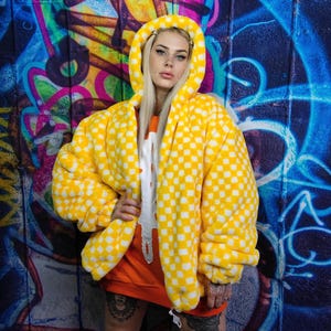 May include: A yellow and white checked hooded jacket. The jacket is oversized and has a soft, plush texture. The person is wearing an orange sweatshirt and a white shirt with a design. The background is a colourful graffiti wall.