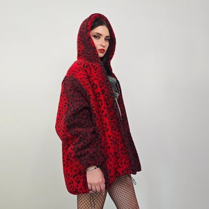 Peut inclure: Une veste en polaire à capuche à imprimé léopard rouge et noir. La veste a une fermeture éclair et est portée par une personne aux cheveux noirs.