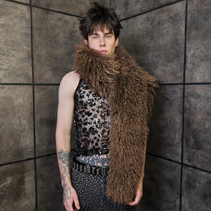Könnte beinhalten: Ein brauner Kunstfellschal über einem schwarzen Pailletten-Tanktop. Das Tanktop hat ein Blumenmuster. Die Person trägt eine Jeans mit Nieten und einen Nietengürtel. Der Hintergrund ist eine graue Kachelwand.