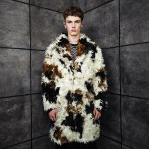 May include: A long, faux fur coat with a pattern of white, brown, and black patches. The coat has a collar and long sleeves. The person is wearing a black top underneath. The background is a gray wall.