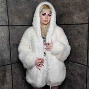 May include: A fluffy, off-white faux fur coat with a large hood. The coat has a relaxed fit and a zip closure. The texture of the fur is visible, creating a soft, voluminous appearance. The coat is a statement piece, perfect for colder weather.