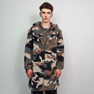 May include: A long, open-front coat in a camouflage pattern featuring shades of green, brown, black, and beige. The coat has a large collar and a soft, textured appearance. The model is wearing a black mesh top and silver jewelry.