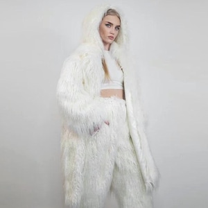May include: A full-length, white faux fur coat with a hood, paired with matching pants. The outfit is made of long, shaggy fur, creating a fluffy, textured appearance. The model is wearing a white crop top and white shoes.