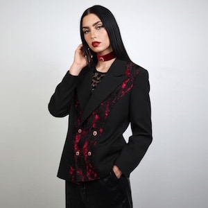 Könnte beinhalten: Schwarzer Blazer mit roten Spitzendetails an Revers und Vorderseite. Der Blazer hat ein doppelreihiges Design mit goldenen Knöpfen. Ein rotes Samt-Choker wird um den Hals getragen. Teil einer Gothic-Modekollektion.