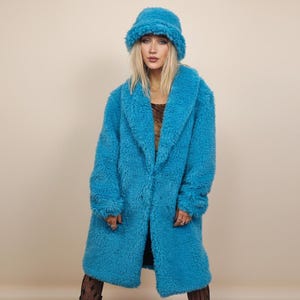May include: A long, turquoise faux fur coat and matching bucket hat. The coat has a wide collar and a knee-length hem. The hat is a soft, textured material. The model is wearing patterned tights and boots.