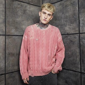 May include: A distressed, light pink cable-knit sweater with long sleeves. The sweater has a worn appearance with intentional rips and a small silver pin near the neckline. The model is wearing black and white patterned pants.
