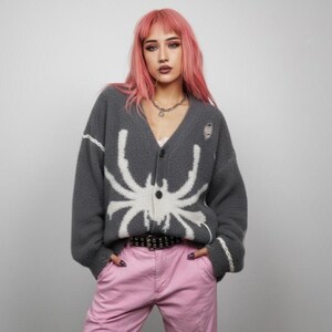 Peut inclure: Un cardigan gris avec un motif d'araignée blanc. Le pull à col en V a des boutons et des détails déchirés. Le modèle porte un pantalon rose et une ceinture noire. Le cardigan est en matière douce.