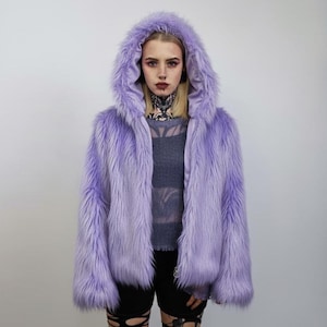 May include: A lavender faux fur hooded jacket. The jacket has a long, shaggy texture and a zip-up front. The model is wearing a gray top and black shorts. The jacket is a vibrant shade of purple.