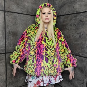 Puede incluir: Chaqueta con capucha y cremallera con un estampado de leopardo vibrante en verde neón, rosa y morado. La chaqueta tiene una textura afelpada y un ajuste holgado. La modelo lleva una falda blanca con un estampado floral.