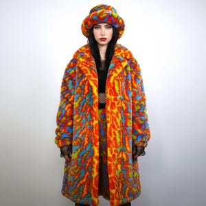 Op de afbeelding: Een felgekleurde imitatiebontjas en bijpassende bucket hat. De lange jas heeft een brede kraag en een opvallend patroon van oranje, geel, blauw en groen. De hoed weerspiegelt het ontwerp van de jas en creëert een opvallend ensemble. De jas is ongeveer 130 cm lang.