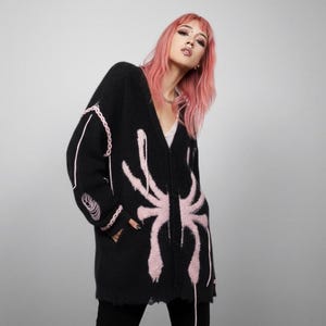 Puede incluir: Cárdigan negro con un diseño de telaraña rosa. El suéter de manga larga tiene cuello en V y dobladillo desgastado. El diseño de telaraña está en la parte delantera del cárdigan. El cárdigan se usa sobre una camiseta blanca.