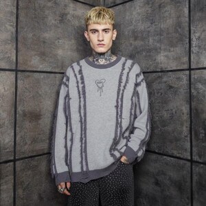 Könnte beinhalten: Ein grauer Pullover mit dunkelgrauen vertikalen Streifen und ausgefransten Kanten. Der Pullover hat eine kleine Herzgrafik mit dem Wort "Rare" auf der Brust. Die Ärmel und der Saum sind ebenfalls dunkelgrau eingefasst.