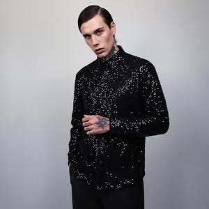 Può includere: Camicia nera abbottonata ricoperta di piccole paillettes riflettenti. La camicia ha un colletto classico e maniche lunghe. Le paillettes sono sparse sul tessuto, creando un effetto scintillante. La camicia è abbinata a pantaloni neri.