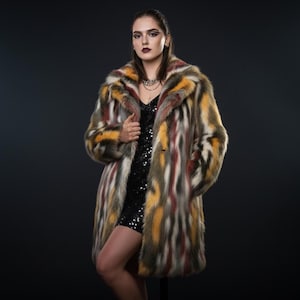 Op de afbeelding: Een veelkleurige imitatiebontjas met verticale strepen in tinten geel, rood, bruin en wit. De jas heeft een klassieke reverskraag en een knielange snit. De persoon draagt een zwarte paillettenjurk.