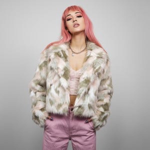 Peut inclure: Veste courte en fausse fourrure à motifs blancs, roses et vert olive. La veste a un large col et des manches longues. Le modèle porte un haut rose et un pantalon rose.