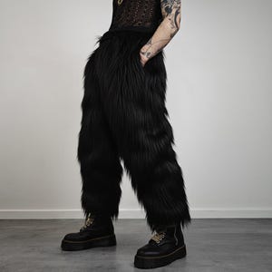 May include: A pair of black faux fur trousers with a wide leg and a shaggy texture. The person is wearing black platform boots. The background is a plain, light grey wall. The trousers are a statement piece.