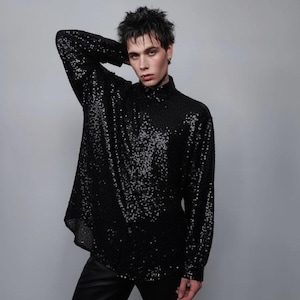 Puede incluir: Camisa negra de manga larga con lentejuelas y cuello alto. La camisa está cubierta de pequeñas lentejuelas reflectantes, creando un efecto brillante. La prenda se usa con pantalones negros. El estilo general es moderno y atrevido.