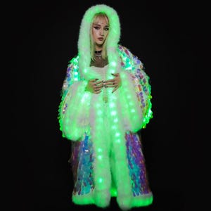 May include: A long, hooded coat with a faux fur trim and iridescent sequins. The coat is illuminated with green LED lights, creating a glowing effect. The coat is worn over a white top and a black background.