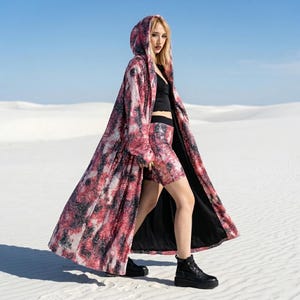 May include: A patterned, hooded coat in red, black, and white, worn over a black top and shorts. The outfit is completed with black boots. The background is a white desert. The coat's length is visible.