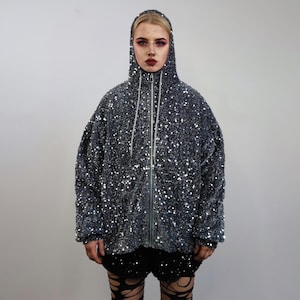 Può includere: Giacca con cappuccio grigio scuro con cerniera a tutta lunghezza, ricoperta di piccole paillettes riflettenti. La giacca ha una vestibilità ampia e un cappuccio. La modella indossa pantaloncini neri.