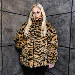 Könnte beinhalten: Eine Kunstfelljacke mit Tigerstreifenmuster in Beige- und Schwarztönen. Die Jacke hat einen hohen Kragen, einen Reißverschluss und lange Ärmel. Die Jacke wird über einer schwarzen Hose getragen.