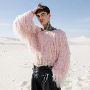 Peut inclure: Un pull rose clair à franges avec des manches longues. Le pull est associé à un pantalon en cuir noir et une ceinture chaîne argentée. Le fond est un paysage désertique avec du sable blanc et un ciel bleu clair.