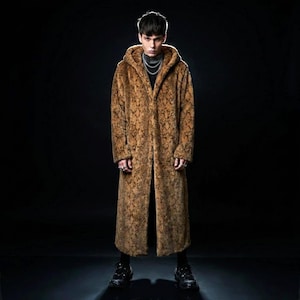 May include: A long, hooded coat with a brown snakeskin pattern. The coat is floor-length and appears to be made of faux fur. The person is wearing a black turtleneck and black shoes. The background is black.
