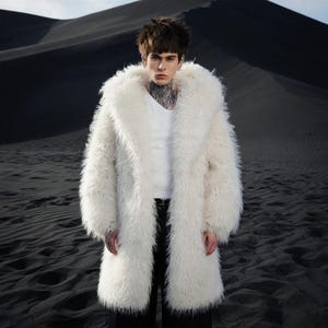 Può includere: Un lungo cappotto in ecopelliccia bianca con un ampio colletto. Il cappotto è indossato sopra una maglietta bianca con scollo a V e pantaloni neri. Lo sfondo presenta dune di sabbia scure sotto un cielo nuvoloso.