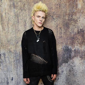 May include: Black distressed knit sweater with open weave detailing. The sweater has a crew neck and long sleeves. The model is wearing a silver necklace and a chain attached to the pants. The background is a textured, neutral-toned wall.