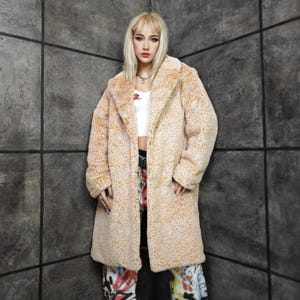 Può includere: Un lungo cappotto in finta pelliccia beige con un motivo leopardato. Il cappotto ha un ampio colletto e un design lungo fino al ginocchio. La modella indossa un top corto bianco e pantaloni colorati in stile graffiti. Lo sfondo è un muro grigio strutturato.