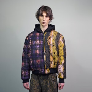 Pode incluir: Uma jaqueta puffer preta com fecho de correr. Um lado da jaqueta apresenta um padrão xadrez em tons de roxo, rosa e preto. O outro lado apresenta um padrão de pele de cobra em tons de amarelo, laranja e preto.