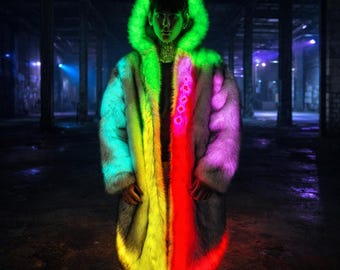May include: A long, open faux fur coat with a vibrant, color-blocked design. The coat features a neon green hood and transitions through shades of teal, yellow, red, and purple. The coat is worn in a dark, industrial setting.