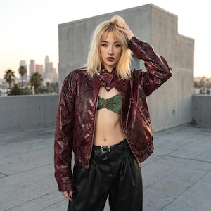 May include: A burgundy faux snakeskin jacket with a unique cutout design, paired with a green top and black pants. The jacket features a zipper closure and a subtle pattern. The outfit is completed with a black belt.
