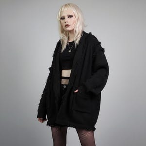 May include: A black, open-front cardigan with a fringed trim and two front pockets. The oversized sweater is made of a thick knit material. The model is wearing a black crop top and fishnet stockings.